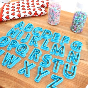 May include: A set of blue plastic alphabet cookie cutters on a wooden surface. The cutters are arranged in alphabetical order. There are two jars of sprinkles in the background.