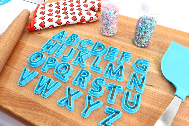 May include: A set of blue plastic alphabet cookie cutters on a wooden surface. The cutters are arranged in alphabetical order. There are two jars of sprinkles in the background.