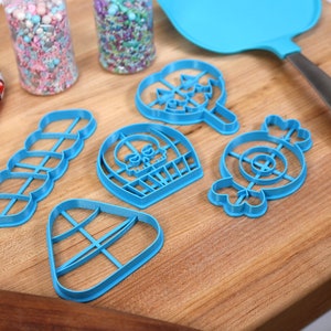 May include: A set of six blue plastic cookie cutters shaped like a skull, a lollipop, a candy cane, a candy corn, a donut, and a string of hearts. The cutters are on a wooden cutting board.
