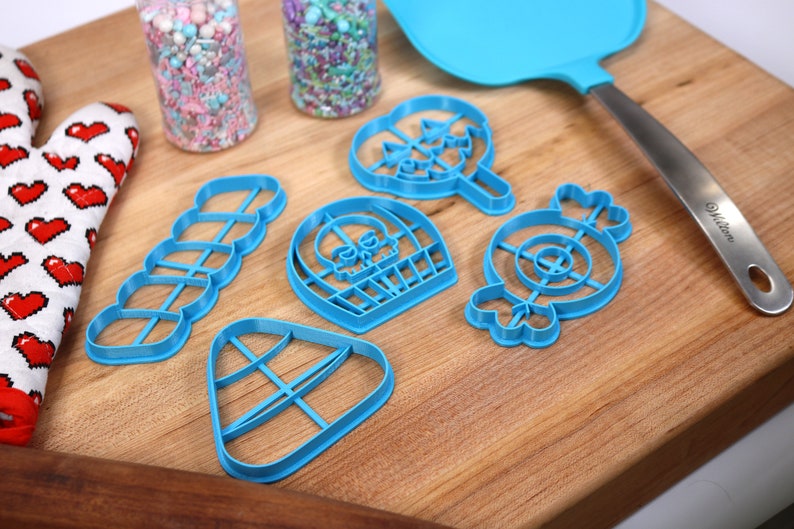 May include: A set of six blue plastic cookie cutters shaped like a skull, a lollipop, a candy cane, a candy corn, a donut, and a string of hearts. The cutters are on a wooden cutting board.