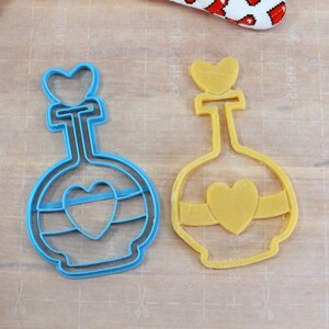 May include: Two cookie cutters in the shape of a bottle with a heart on top. The bottle on the left is blue and the bottle on the right is yellow.