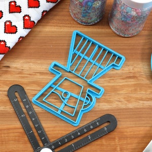 May include: A blue plastic cookie cutter shaped like a coffee grinder. The cookie cutter is on a wooden surface with a ruler and a spatula.