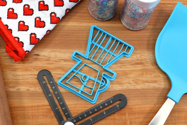 May include: A blue plastic cookie cutter shaped like a coffee grinder. The cookie cutter is on a wooden surface with a ruler and a spatula.