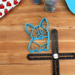 Pu&ograve; includere: Tagliapasta in plastica blu a forma di corgi di cartone animato. Il tagliapasta &egrave; su una superficie di legno con un righello nero.