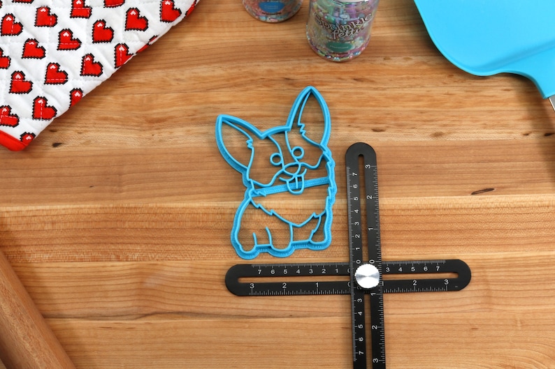 Pu&ograve; includere: Tagliapasta in plastica blu a forma di corgi di cartone animato. Il tagliapasta &egrave; su una superficie di legno con un righello nero.