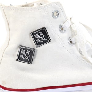 May include: White high-top canvas sneakers with two silver metal square-shaped pins with a design of three swords. The pins are attached to the side of the shoe.