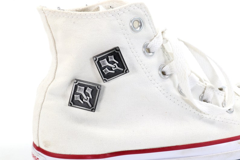 May include: White high-top canvas sneakers with two silver metal square-shaped pins with a design of three swords. The pins are attached to the side of the shoe.