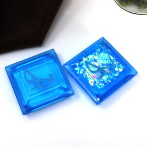 May include: Two square blue glass tiles with a white design. One tile has a white design of a rocking chair, the other tile has a white design of a butterfly with iridescent glitter inside.