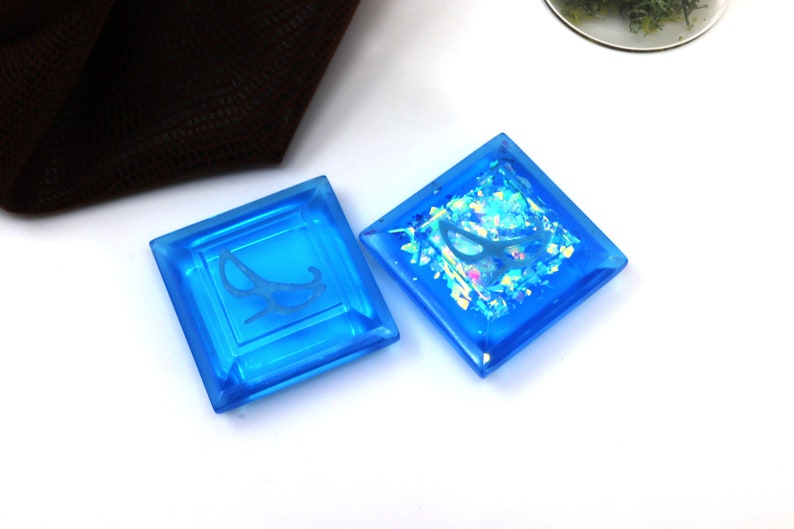 May include: Two square blue glass tiles with a white design. One tile has a white design of a rocking chair, the other tile has a white design of a butterfly with iridescent glitter inside.