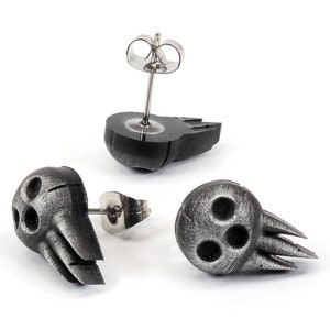 May include: Three silver skull earrings with a textured finish. The earrings are in the shape of a skull with three holes for eyes and a mouth. The earrings are on a white background.