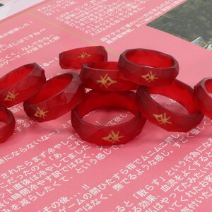 May include: A group of eight red resin rings with a gold design. The rings are stacked on top of each other and are arranged in a circle.