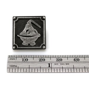 May include: A silver metal pin with a square frame and a stylized image of a creature inside. The pin is about 2 cm wide.