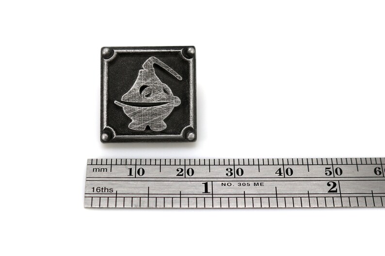 May include: A silver metal pin with a square frame and a stylized image of a creature inside. The pin is about 2 cm wide.
