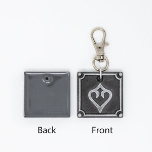 May include: A gray metal keychain with a silver heart design on the front and a plain gray back. The keychain has a silver metal clasp.