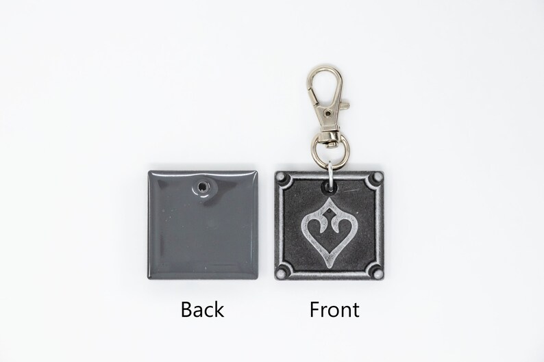 May include: A gray metal keychain with a silver heart design on the front and a plain gray back. The keychain has a silver metal clasp.