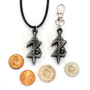 May include: A silver pendant necklace and keychain featuring a stylized sword with a Z design. The pendant is on a black cord and the keychain has a silver clip. Both are shown next to US pennies and a British pound coin for scale.