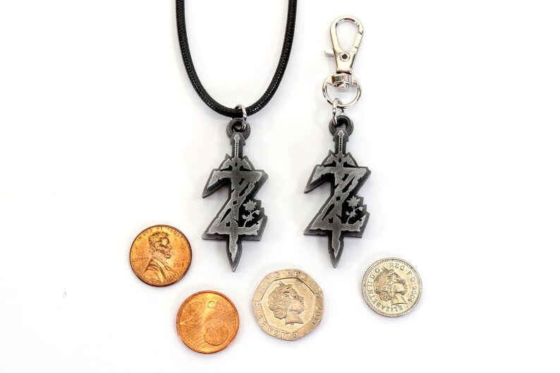 May include: A silver pendant necklace and keychain featuring a stylized sword with a Z design. The pendant is on a black cord and the keychain has a silver clip. Both are shown next to US pennies and a British pound coin for scale.