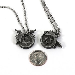K&ouml;nnte beinhalten: Zwei silberne Halsketten mit Anh&auml;ngern, die das alchemistische Symbol f&uuml;r Transmutation zeigen, einen Kreis mit einem Dreieck darin und eine stilisierte gefl&uuml;gelte Kreatur. Die Anh&auml;nger sind etwa so gro&szlig; wie eine deutsche Zehn-Cent-M&uuml;nze.