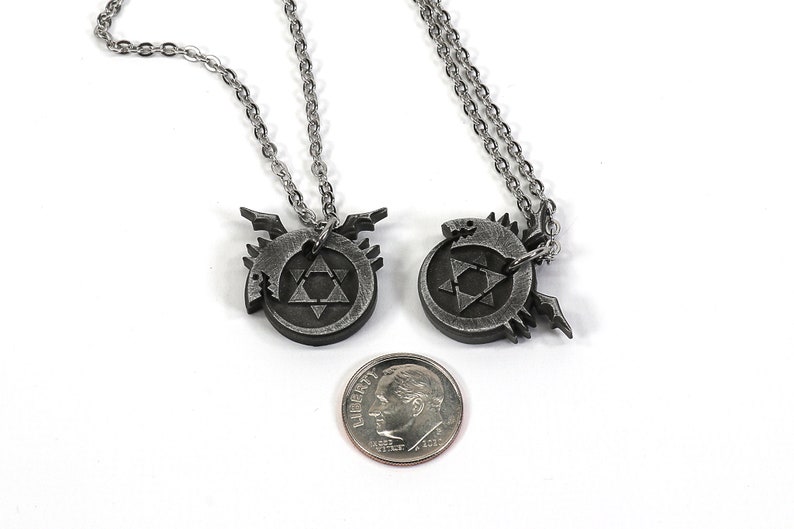 K&ouml;nnte beinhalten: Zwei silberne Halsketten mit Anh&auml;ngern, die das alchemistische Symbol f&uuml;r Transmutation zeigen, einen Kreis mit einem Dreieck darin und eine stilisierte gefl&uuml;gelte Kreatur. Die Anh&auml;nger sind etwa so gro&szlig; wie eine deutsche Zehn-Cent-M&uuml;nze.
