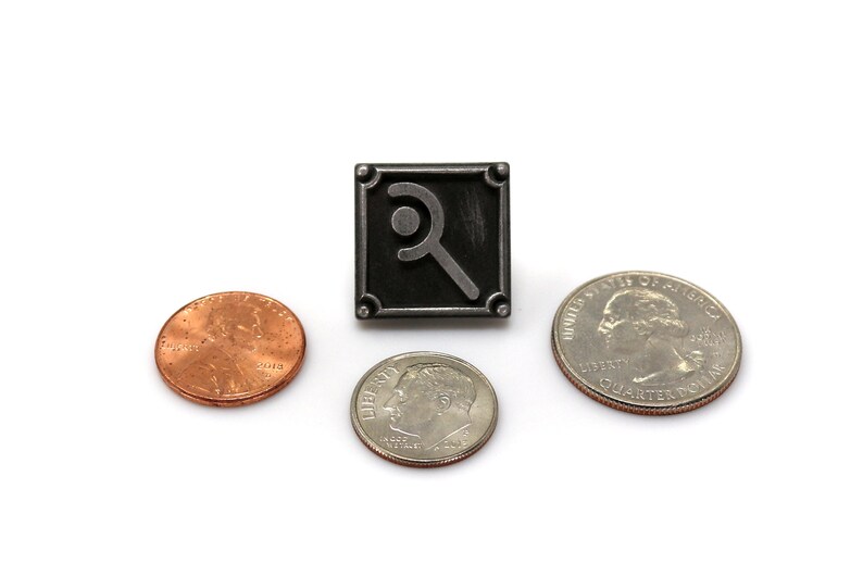 May include: A square, dark metal pin with a stylized magnifying glass design, flanked by a penny, a dime, and a quarter. The pin has rounded corners and a raised border. The coins are arranged around the pin on a white background.