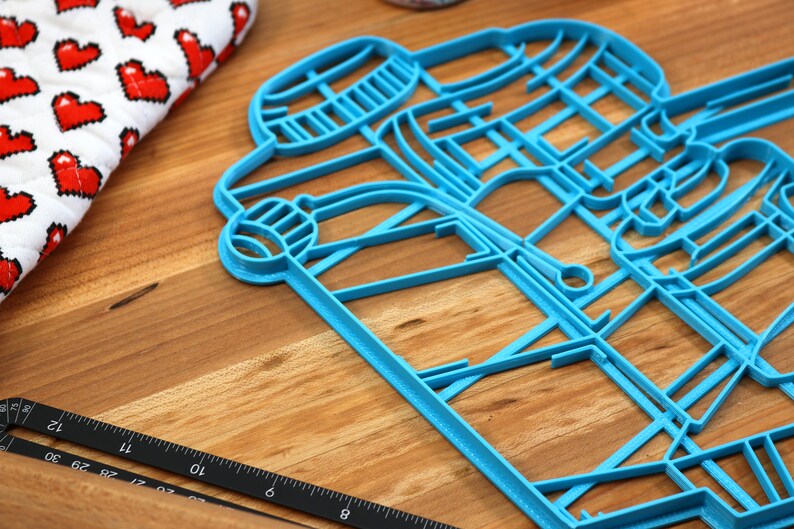 May include: A blue plastic cookie cutter shaped like a pair of coffee cups. The cookie cutter is on a wooden surface.
