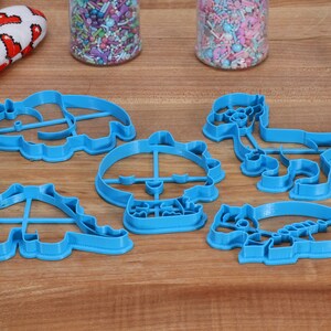 May include: A set of six blue plastic dinosaur-shaped cookie cutters. The cutters are arranged on a wooden surface.