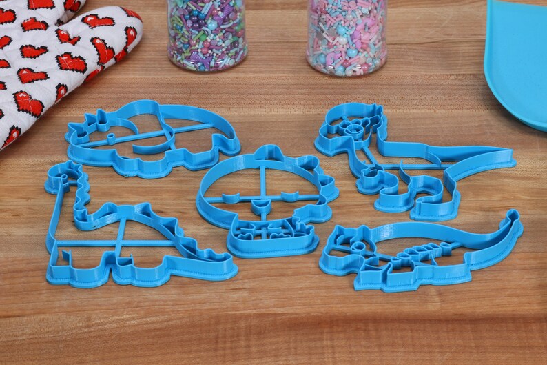 May include: A set of six blue plastic dinosaur-shaped cookie cutters. The cutters are arranged on a wooden surface.