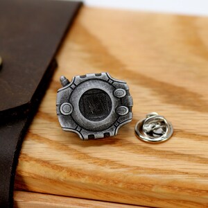 May include: A silver-colored metal pin shaped like a digital device with a circular screen and buttons. The pin has a silver-colored metal pin back.