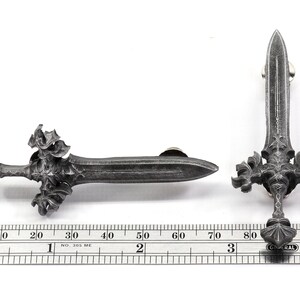 May include: A silver-colored metal sword pin with intricate details. The sword has a pointed tip and a cross-shaped hilt with decorative flourishes. The pin is approximately 3 inches long.