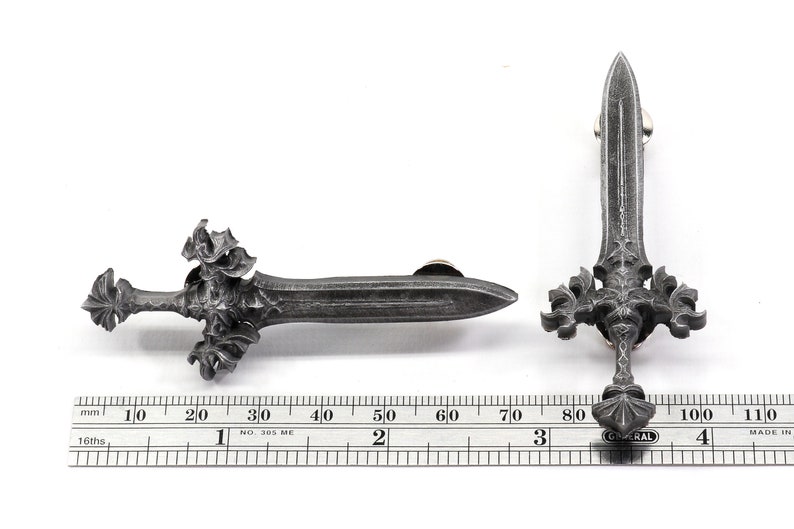 May include: A silver-colored metal sword pin with intricate details. The sword has a pointed tip and a cross-shaped hilt with decorative flourishes. The pin is approximately 3 inches long.