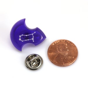 May include: A purple resin pin with a white outline of the Gemini constellation. The pin is next to a US penny for size comparison.