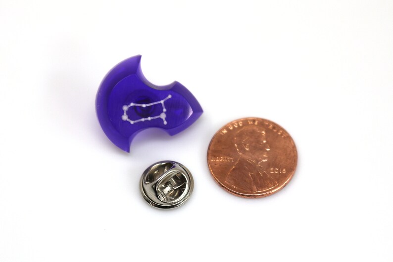 May include: A purple resin pin with a white outline of the Gemini constellation. The pin is next to a US penny for size comparison.