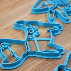 Puede incluir: Un juego de cortadores de galletas de pl&aacute;stico azul con forma de varios dise&ntilde;os navide&ntilde;os. Los cortadores est&aacute;n sobre una superficie de madera.