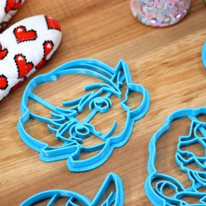 May include: Four blue plastic cookie cutters shaped like cartoon characters. The cutters are on a wooden surface.