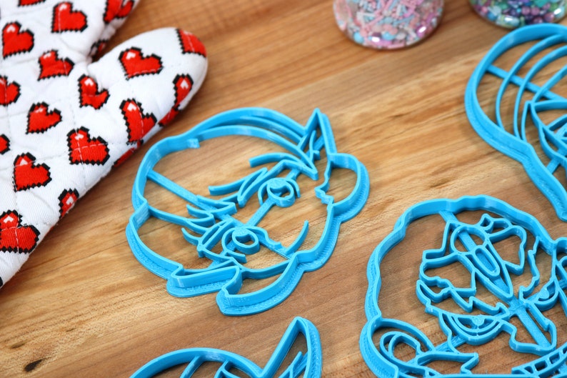 May include: Four blue plastic cookie cutters shaped like cartoon characters. The cutters are on a wooden surface.