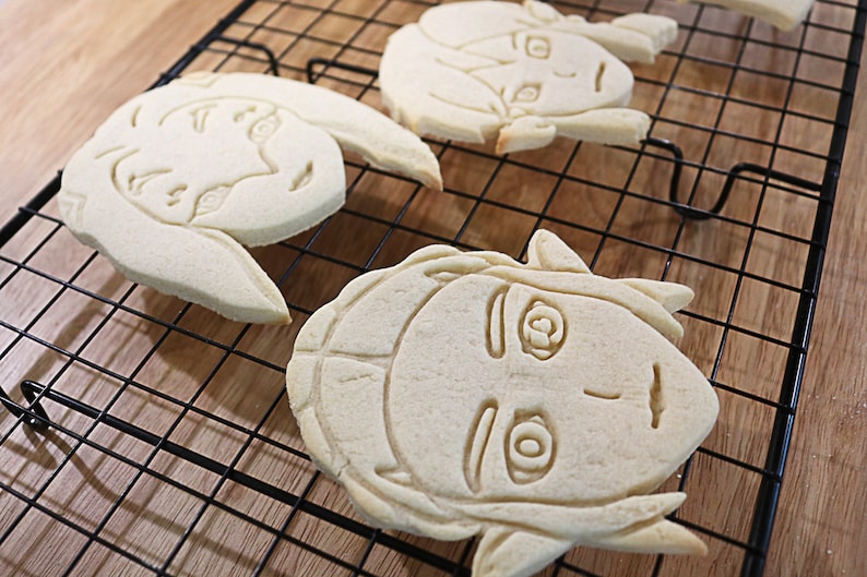 May include: Three sugar cookies decorated with icing in the shape of cartoon faces. The cookies are on a black wire cooling rack.