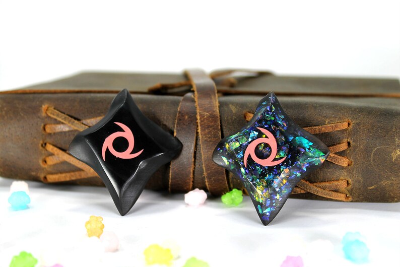 Pu&ograve; includere: Due stelle ninja nere a forma di stella con un design a spirale rosa. Una stella &egrave; realizzata in un materiale nero liscio, mentre l'altra &egrave; realizzata in un materiale nero scintillante con riflessi blu e verdi iridescenti.