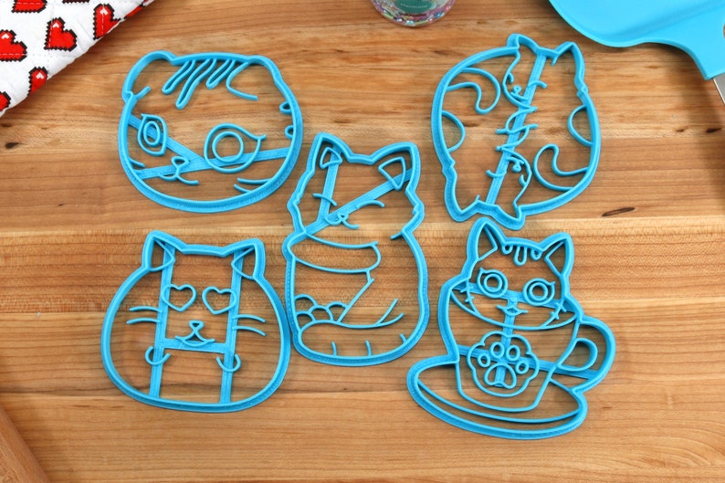 May include: Five blue plastic cookie cutters shaped like cats. The cutters feature different cat designs, including a cat with hearts for eyes, a cat in a cup, and a cat with a curled tail.