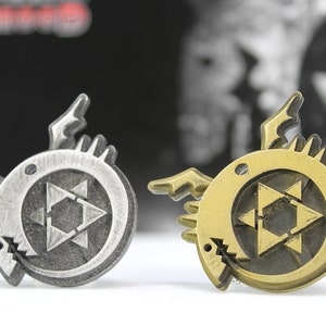 May include: Two metal pins featuring a stylized black star with a circle around it. The star is surrounded by a circle with a creature's head on each side. One pin is silver and the other is gold.