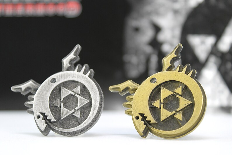 May include: Two metal pins featuring a stylized black star with a circle around it. The star is surrounded by a circle with a creature's head on each side. One pin is silver and the other is gold.
