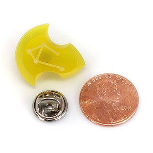 May include: Yellow enamel pin with a white outline of the Libra constellation. The pin is about the size of a US penny.