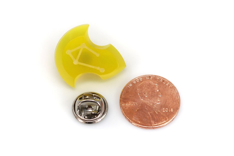 May include: Yellow enamel pin with a white outline of the Libra constellation. The pin is about the size of a US penny.
