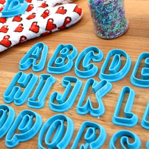 May include: A set of blue plastic alphabet cookie cutters on a wooden surface. The cutters are arranged in alphabetical order from A to Z.