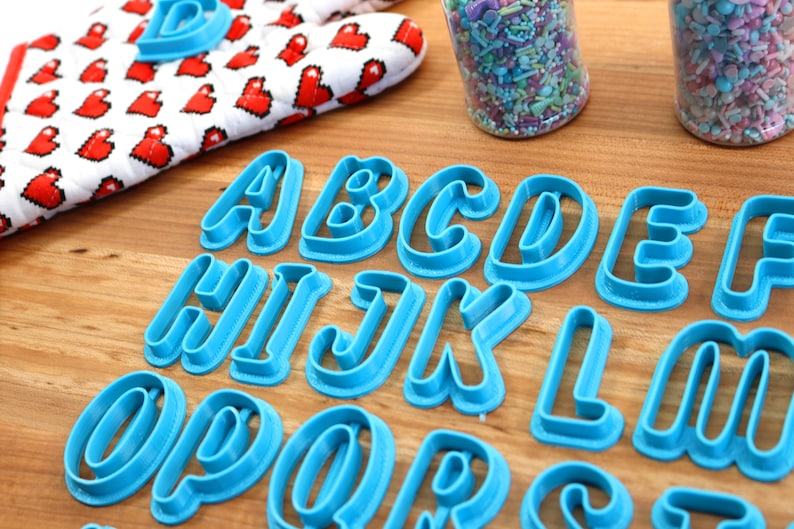 May include: A set of blue plastic alphabet cookie cutters on a wooden surface. The cutters are arranged in alphabetical order from A to Z.