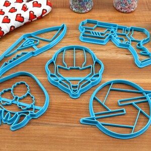 May include: A set of five blue plastic cookie cutters shaped like a space helmet, a gun, a creature, a knife, and a logo.