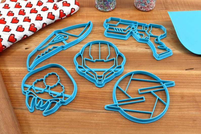 May include: A set of five blue plastic cookie cutters shaped like a space helmet, a gun, a creature, a knife, and a logo.