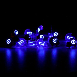May include: A group of small, clear, glowing purple objects with white symbols on them. The objects are scattered on a black surface.