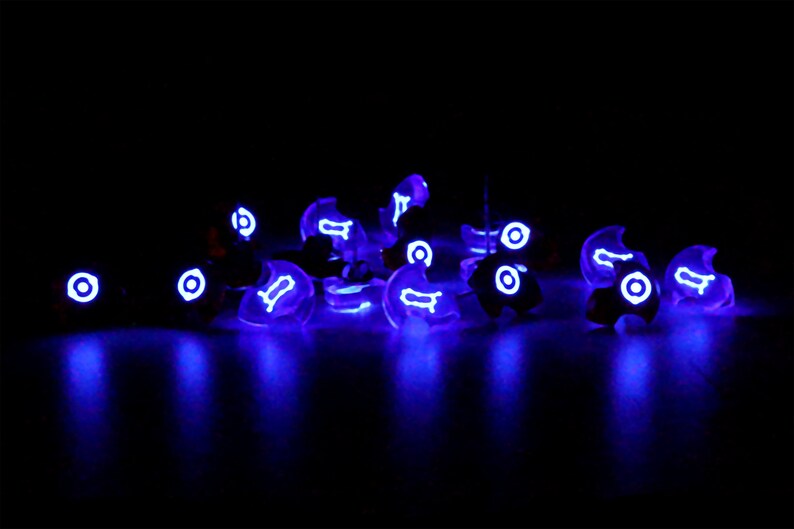 May include: A group of small, clear, glowing purple objects with white symbols on them. The objects are scattered on a black surface.