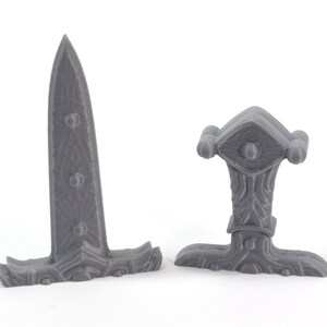 May include: Two gray plastic fantasy sword hilt designs. One is a straight hilt with a decorative eye design. The other is a curved hilt with a decorative design.