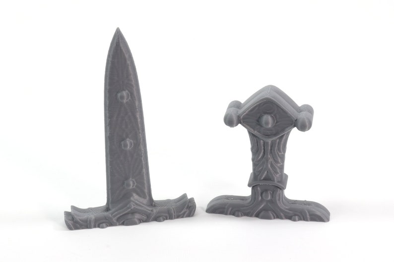 May include: Two gray plastic fantasy sword hilt designs. One is a straight hilt with a decorative eye design. The other is a curved hilt with a decorative design.
